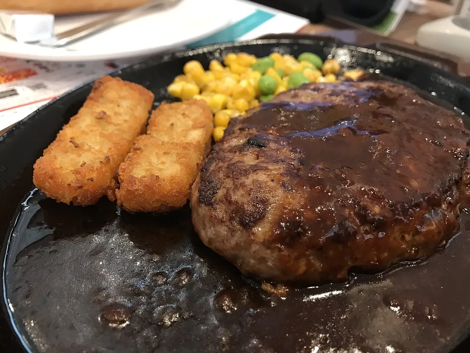 ハンブルクステーキの実写写真（日本の洋食ハンバーグ(ハンブルクステーキ)=挽肉成形のデミ/ソース掛け鉄板ハンバーグの現行完成皿。ファミレス(ガスト)盛付け）