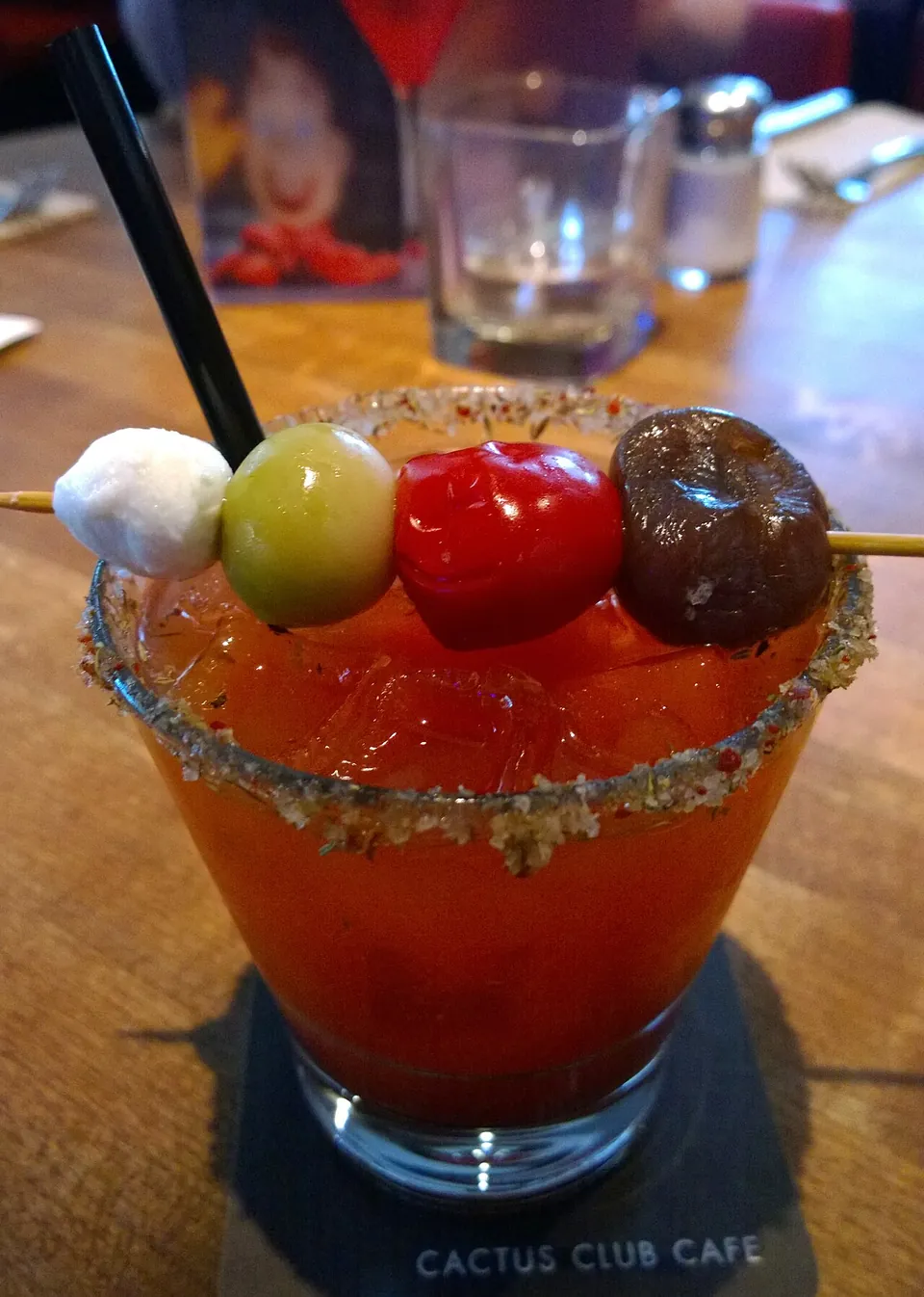 シーザー(ブラッディ・シーザー)の実写写真（カナダのブラッディ・シーザー(Cactus Club Cafe)。クラマト系トマトジュースの赤・氷入り・縁までなみなみ注がれた満杯グラス(飲みかけでない)・ハーブ入りリム塩・ガーニッシュ串。却下img#422『飲みかけなのが良くない』への対応=注ぎたて満杯の別画像。）